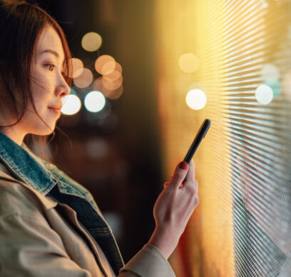 Young Asian woman using smartphone against illuminated digital display in city street at night. Blockchain technology. Trading cryptocurrency. NFT (Non-Fungible Token) investment.